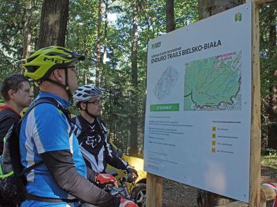 Zdjęcie przedstawiające mężczyzn przy tablicy informacyjnej o górskich ścieżkach enduro wśród zieleni.