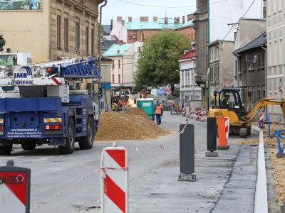 Zdjęcie przedstawiające plac budowy z koparką i robotnikami w kaskach, prowadzącymi prace ziemne w otoczeniu bloków mieszkalnych.