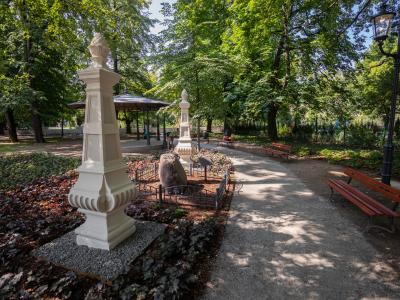 Zdjęcie przedstawiające zacienioną alejkę parkową z pomnikami, ławkami i altaną.