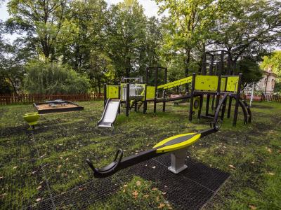 Zdjęcie przedstawiające plac zabaw z huśtawką, zjeżdżalnią i konstrukcjami do wspinaczki w otoczeniu drzew.