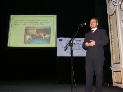 zdjęcie mężczyzny w garniturze przemawiającego na scenie obok prezentacji multimedialnej.