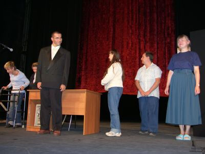 zdjęcie grupa osób stojącej na scenie teatralnej przed biurkiem, na tle czerwonej kurtyny