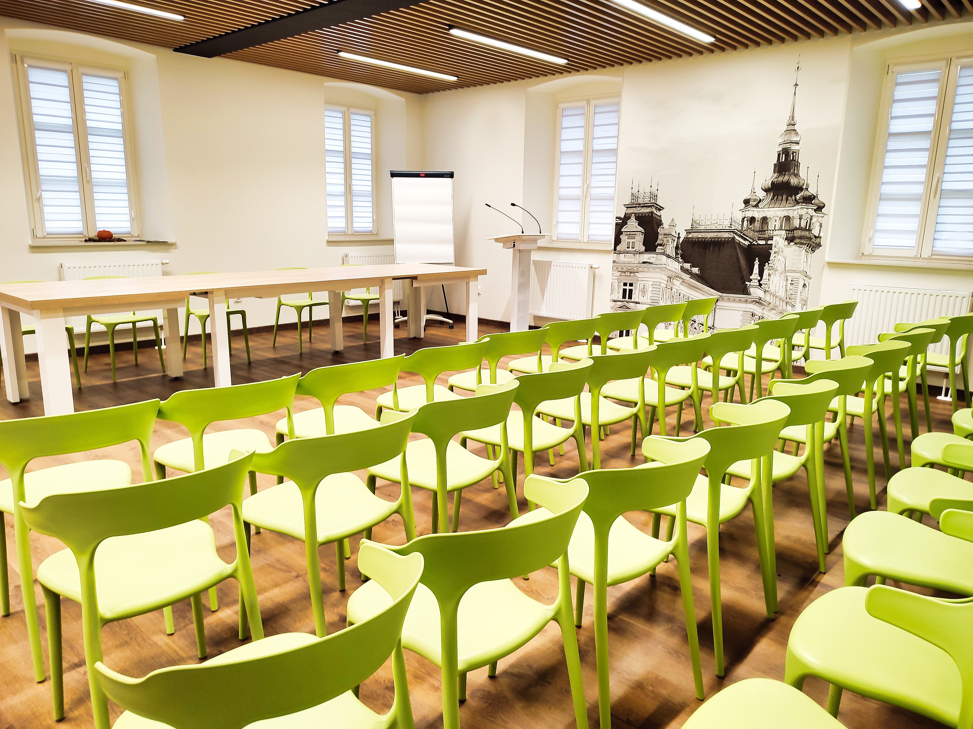 zdjęcie przedstawiające sale ze stołem, czterema rzędami krzeseł, flipchartem oraz mównicą