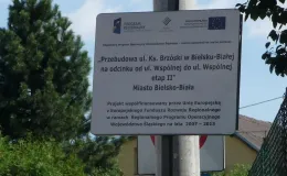 Przebudowa ul. Ks. Brzóski w Bielsku-Białej - na zdjęciu tablica informacyjna, budynek, znak drogowy oraz zieleń