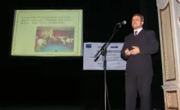 zdjęcie mężczyzny w garniturze przemawiającego na scenie obok prezentacji multimedialnej.