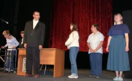 zdjęcie grupa osób stojącej na scenie teatralnej przed biurkiem, na tle czerwonej kurtyny