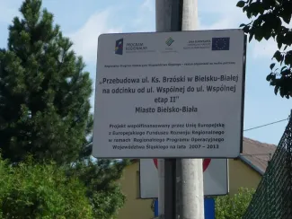 Przebudowa ul. Ks. Brzóski w Bielsku-Białej - na zdjęciu tablica informacyjna, budynek, znak drogowy oraz zieleń