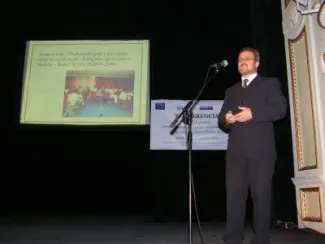 zdjęcie mężczyzny w garniturze przemawiającego na scenie obok prezentacji multimedialnej.