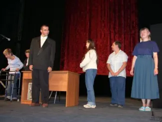 zdjęcie grupa osób stojącej na scenie teatralnej przed biurkiem, na tle czerwonej kurtyny