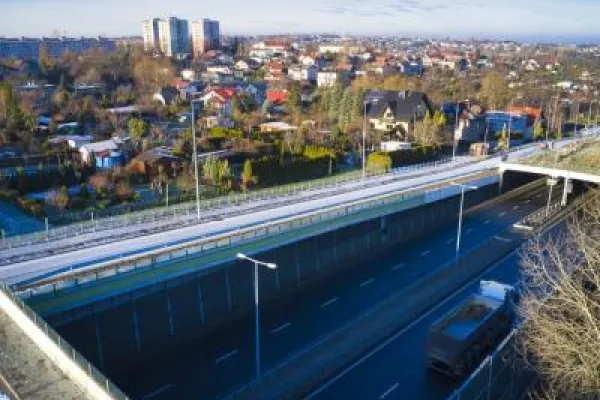Zdjęcie przedstawiające drogę ekspresową w wykopie z wiaduktem i zabudową mieszkalną w tle.