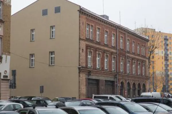 Zdjęcie przedstawiające budynek z ceglaną elewacją przy parkingu pełnym samochodów.