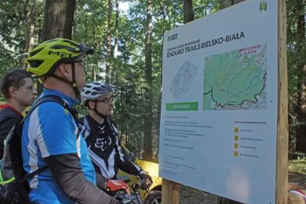 Zdjęcie przedstawiające mężczyzn przy tablicy informacyjnej o górskich ścieżkach enduro wśród zieleni.