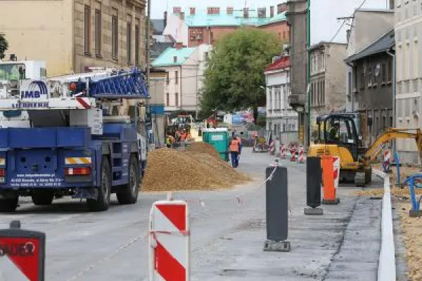 Zdjęcie przedstawiające plac budowy z koparką i robotnikami w kaskach, prowadzącymi prace ziemne w otoczeniu bloków mieszkalnych.