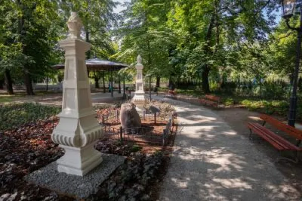 Zdjęcie przedstawiające zacienioną alejkę parkową z pomnikami, ławkami i altaną.