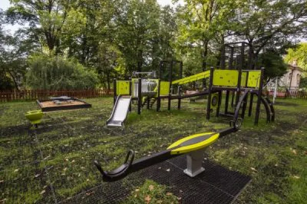 Zdjęcie przedstawiające plac zabaw z huśtawką, zjeżdżalnią i konstrukcjami do wspinaczki w otoczeniu drzew.