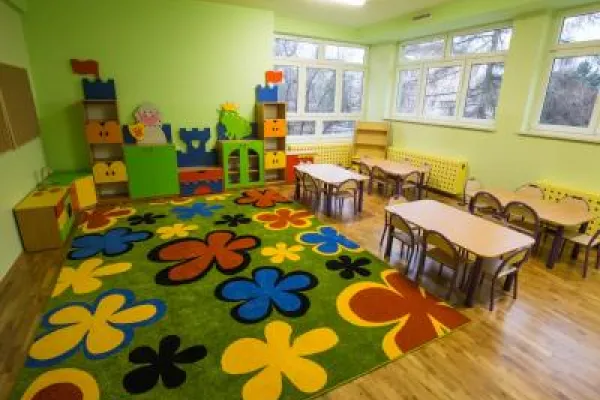 Zdjęcie przedstawiające salę przedszkolną z dywanem w kwiaty oraz małymi stolikami i krzesłami.